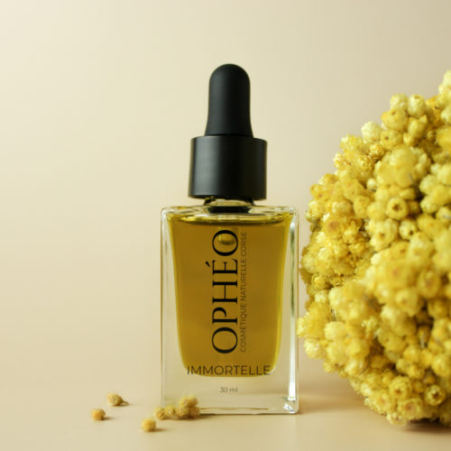 Flacon en verre avec pipette compte-goutte, contenant de un macérat d'immortelle corse pour le visage. Devant le flacon se trouvent des fleurs d'immortelle corses.